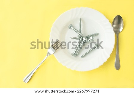 in a white plate lie gray metal bolts with kitchen utensils fork and spoon on a yellow background