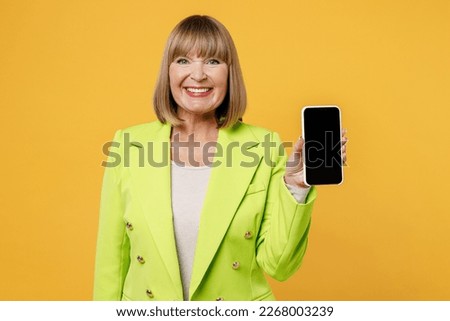 Elderly happy woman 50s years old wearing green jacket white t-shirt hold in hand use mobile cell phone with blank screen workspace area copy space isolated on plain yellow background studio portrait