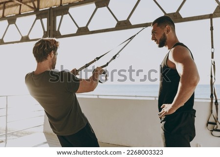 Young sporty strong athletic toned fit sportsman man in sports clothes warm up suspended training TRX work out with instructor at sunrise sun dawn over sea beach seaside outdoor in summer day morning. Royalty-Free Stock Photo #2268003323