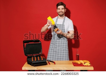 Young smiling housewife housekeeper chef cook baker man in grey apron work at table with grill kitchenware make hotdog add mustard isolated on plain red background studio Process cooking food concept Royalty-Free Stock Photo #2268003325