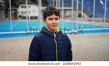 One pensive young boy stands outside in city park by basketball court with hands in jacket pocket. Thoughtful expression