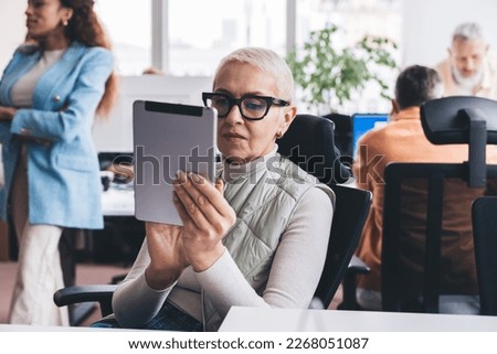 Mature woman wearing eyeglasses sitting on comfortable office chair and looking at screen while working with tablet in hands on project