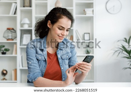 Young curly brunette sitting at the table uses a mobile phone. Surfs the Internet, communicates in instant messengers on his smartphone. Online communication, smartphone in human life. Royalty-Free Stock Photo #2268051519
