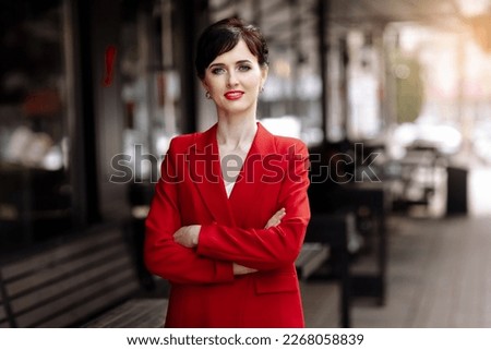 Smiling business woman dressed red jacket and red lipstick standing outdoor near glass corporate office building hands folded. Serious caucasian female business person portrait on city street Royalty-Free Stock Photo #2268058839