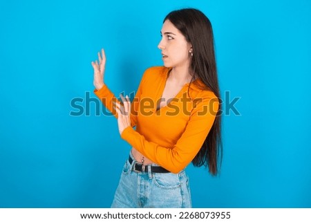 Displeased young woman wearing orange crop top over blue studio background keeps hands towards empty space and asks not come closer sees something unpleasant
