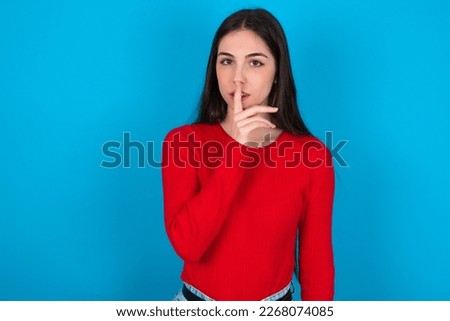 young brunette woman wearing red shirt over white studio background makes silence gesture, keeps finger over lips. Silence and secret concept.