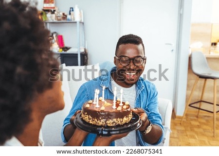 Happy millennial couple celebrating birthday with festive cake at home, full length. Cheerful African American  guy greeting excited girlfriend with b-day, enjoying holiday in living room