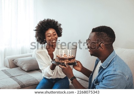 Happy millennial couple celebrating birthday with festive cake at home, full length. Cheerful African American  guy greeting excited girlfriend with b-day, enjoying holiday in living room