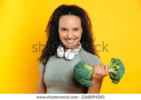 Happy excited cheerful mixed race young sporty woman with curly hair, with headphones on shoulders, holding imaginary broccoli kettlebell, standing on isolated yellow background, smiles, having fun