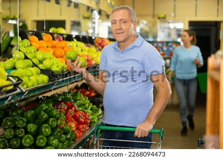 in supermarket, elderly man carefully selects green apples for fruit salad and puts them in shopping cart