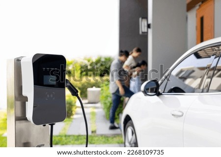 Focus closeup electric vehicle recharging battery from home electric charging station with blurred family in background. Renewable clean energy car for progressive eco awareness lifestyle concept. Royalty-Free Stock Photo #2268109735