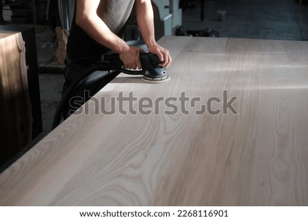 young man builder carpenter grinds a wooden table with a grinding machine in the backyard. A construction worker using a cordless power sander to sand a massive wooden table top Royalty-Free Stock Photo #2268116901