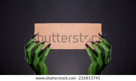 Green monster hands with black nails holding empty piece of cardboard, Halloween theme  