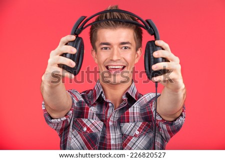 picture of young smiling man offering headphones. guy holding and giving headphones on red background