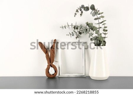 Blank frame, vase with eucalyptus branches and Easter egg on table near white wall