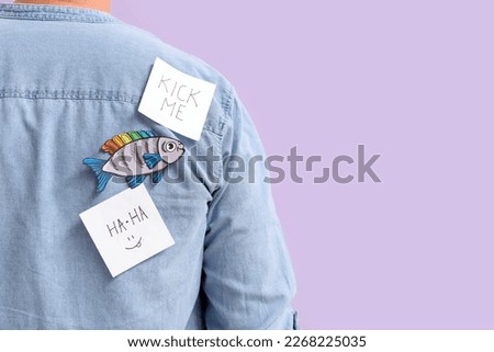 Mature man with sticky papers and fish on lilac background, back view. April Fools' Day celebration Royalty-Free Stock Photo #2268225035