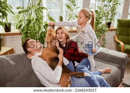 Portrait of beautiful young man and woman, playing with child and don at home on sofa. Happiness, positive vibes and love. Concept of relationship, family, parenthood, childhood, animal life