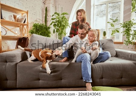Lovely parents, couple, family playing with their dog in living room on warm sunny day at home. Relaxation and happiness. Concept of relationship, family, parenthood, childhood, animal life