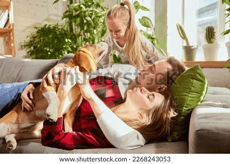 Happy family cuddling their dog beagle, having fun together in living room. Young european couple on the sofa at home, relaxing. Concept of relationship, family, parenthood, childhood, animal life