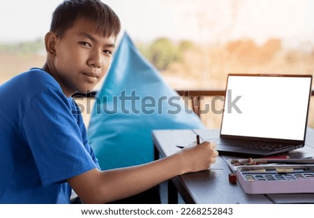 asian boy spends his freetimes with drawing, coloring, painting and sketching pictures in front of labtop on table in the middle of living room outside his house, soft and selective focus.
