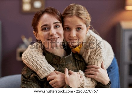 Portrait of smiling military woman with daughter embracing and looking at camera at home