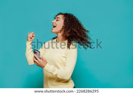 Side view of young cheerful brunette woman, holds smartphone, moving curly hair in the air, celebrates victory rising clenched fist. Studio portrait isolated over blue background.