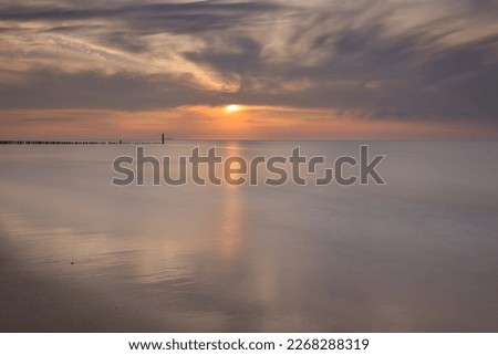 Horizontal view on row of pile heads leading into water. North sea beach during sunset with sun and clouds at the sky. Calm seascape of breakwaters in Zeeland with copy space, slow shutter speed
