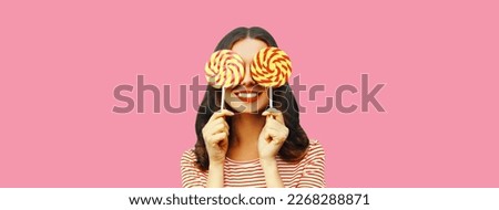 Portrait of cheerful smiling young woman covering her eyes with lollipop on pink background