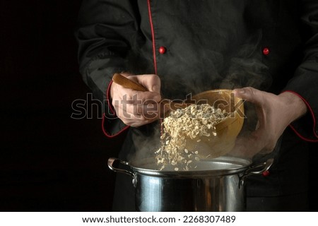 The chef is spooning Rolled oats into the pan. Black space for recipe. The idea of cooking delicious porridge with oatmeal in the kitchen of a restaurant. Royalty-Free Stock Photo #2268307489