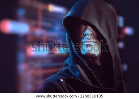 Portrait of a focused man with a confident look, dressed in a black robe with a hood on his head against the background of digital holograms. Cyber theme. Bitcoin, cryptocurrency. Copy space. 