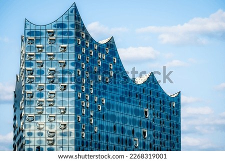 Stunning closeup of the famous glossy glass facade of the iconic Elbphilharmonie concert hall landmark in Hamburg, showcasing its modern, wave-like design and reflecting the sky above in sunlight. Royalty-Free Stock Photo #2268319001