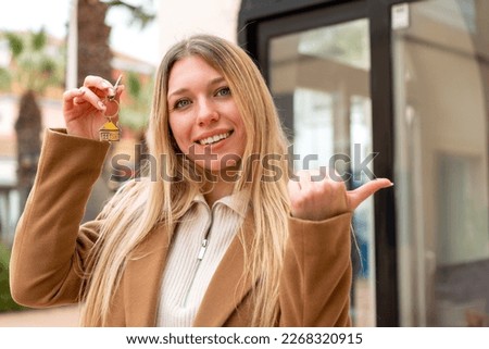 Young pretty blonde woman holding home keys at outdoors pointing to the side to present a product