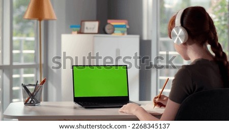 Back view of girl teenager having online lesson using laptop and headphones at home. Rear view of redhead teen pupil study online at home watching webinar on computer. Green screen. High quality photo Royalty-Free Stock Photo #2268345137