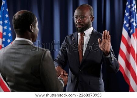 Serious young African American male president pronouncing oath of office with his right hand on Holy Bible while keeping open left one Royalty-Free Stock Photo #2268373955