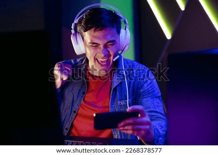 Male video game player cheering and celebrating as he streams a live broadcast of a game on his smartphone. Young man joining an online audience in watching a virtual gameplay on a mobile app. Royalty-Free Stock Photo #2268378577