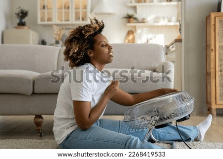 Millennial African American girl sitting on floor in living room feeling hot during extreme summer heat, using electric fan at home, young unhappy black women dealing with summertime hot flashes Royalty-Free Stock Photo #2268419533