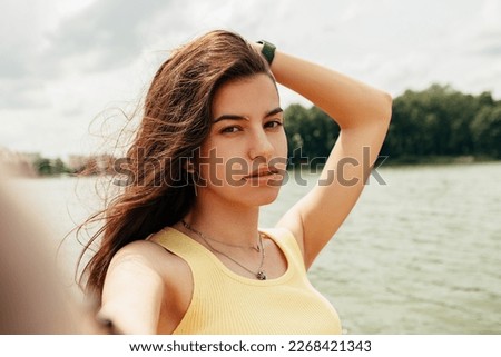Woman standing smiling laughing near river sea lake, take picture, make selfie hold camera. Female on beach island. Girl tourist vacation, weekend relaxing resting resort. Lady go on shore, sunbathing