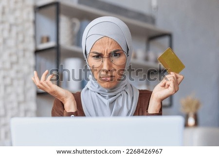 Close-up photo. Credit card problems. Worried young Muslim woman in hijab sitting in front of laptop. He looks at the monitor in shock, spreads his hands.