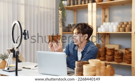 Asian young adult man is giving a presentation about a earthenware bowl by live streaming online at home. Active male merchant is using his smartphone to show details of goods to online customers.