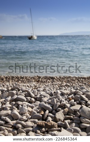 White sandy beach with rough high sea, sail boat and sunny blue sky in the background, selective focus on stones, romantic blue toned, high resolution photo