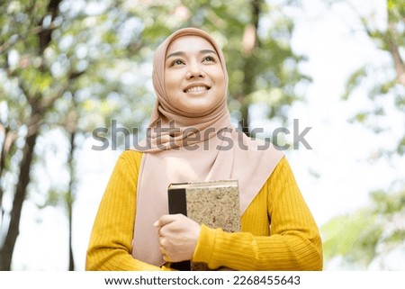 Happy cheerful Asian beautiful female muslim student holding a book and looking and smiling to camera. Modern muslim lifestyles and diversity concept.