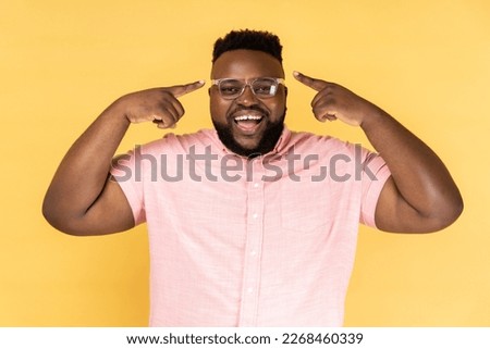 Portrait of delighted satisfied man wearing pink shirt standing, pointing at his new optical spectacles, being happy to see better, smiling happily. Indoor studio shot isolated on yellow background.