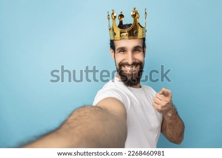Portrait of smiling man with beard wearing white T-shirt and gold crown, pointing at camera, POV, point of view of photo, expressing happiness. Indoor studio shot isolated on blue background.