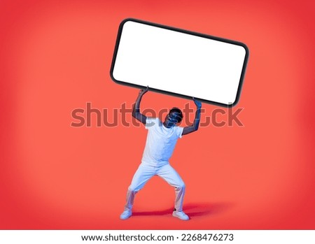 Amazed excited happy handsome young black guy in white lifting up huge cell phone with white empty screen on red background in neon light, mockup for online offer, mobile app advertisement