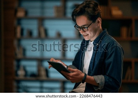 Happy millennial man in glasses use tablet, planning or visualizing future success. Smiling young male working on pad gadget, dreaming or thinking at dark office. Business vision concept.