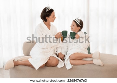 Mom And Little Daughter Wearing Bathrobes And Toy Crowns Relaxing On Couch After Spa Day At Home, Cute Girl And Her Mother Drinking Hot Chocolate And Enjoying Time Together, Copy Space