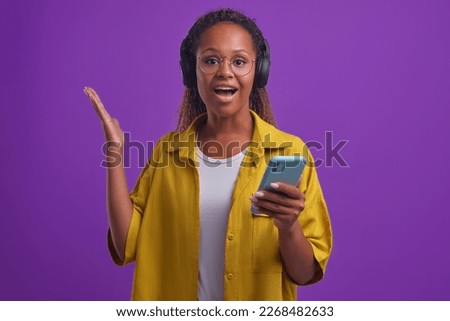 Young admiring ethnic African American woman dressed in casual clothes with mobile phone in hand experiencing wow emotions after learning about cool sale in mall stands posing in lilac studio