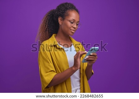 Young attractive African American woman millennial with phone in hands spends all free time on smartphone suffering from gadget addiction and in need of psychological help stands on lilac background Royalty-Free Stock Photo #2268482655