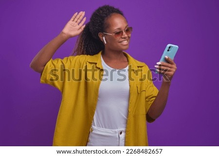 Young sociable African American woman blogger waving hand saying hello to subscribers shooting short video on front camera of mobile phone for vlog stands on lilac background. Shorts, history concept