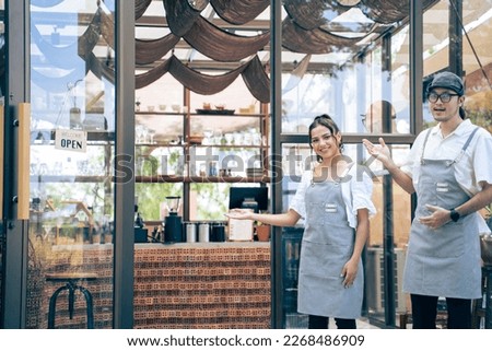 Portrait of Caucasian man and woman waiter looking at camera in cafe. Attractive young entrepreneur barista people wearing apron turning "welcome we are open" sign to open urban coffeehouse shop.